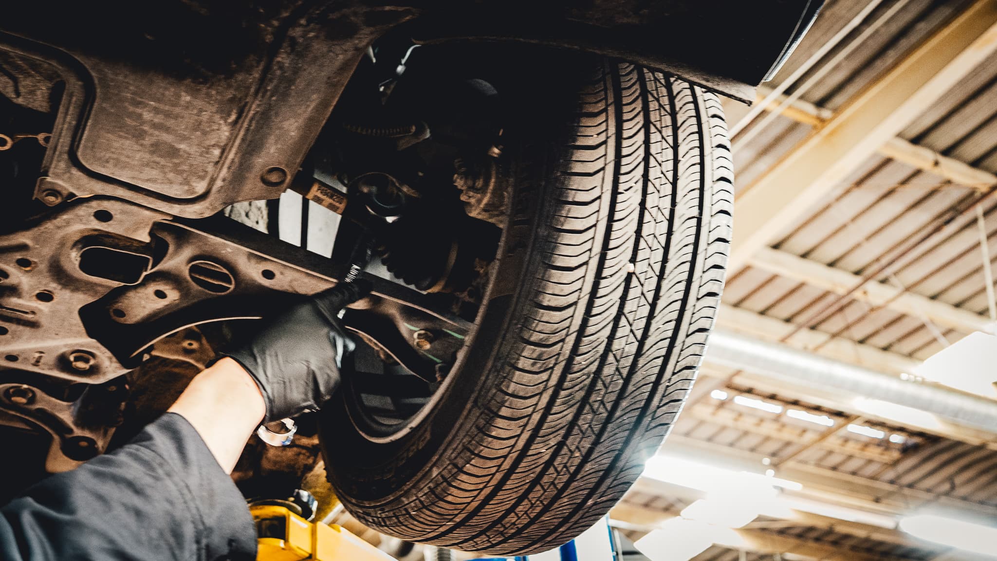 Technician inspecting suspension and tire components under a raised vehicle.